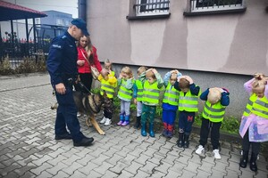 Policjant i pies służbowy. Dzieci w kamizelkach odblaskowych stojące przed budynkiem.