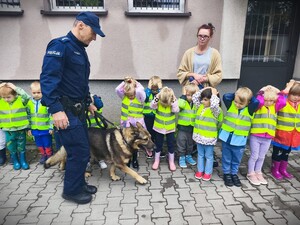 Policjant i pies służbowy. Dzieci w kamizelkach odblaskowych stojące przed budynkiem.