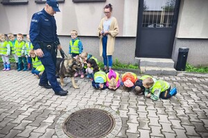Policjant i pies służbowy. Dzieci w kamizelkach odblaskowych stojące przed budynkiem.